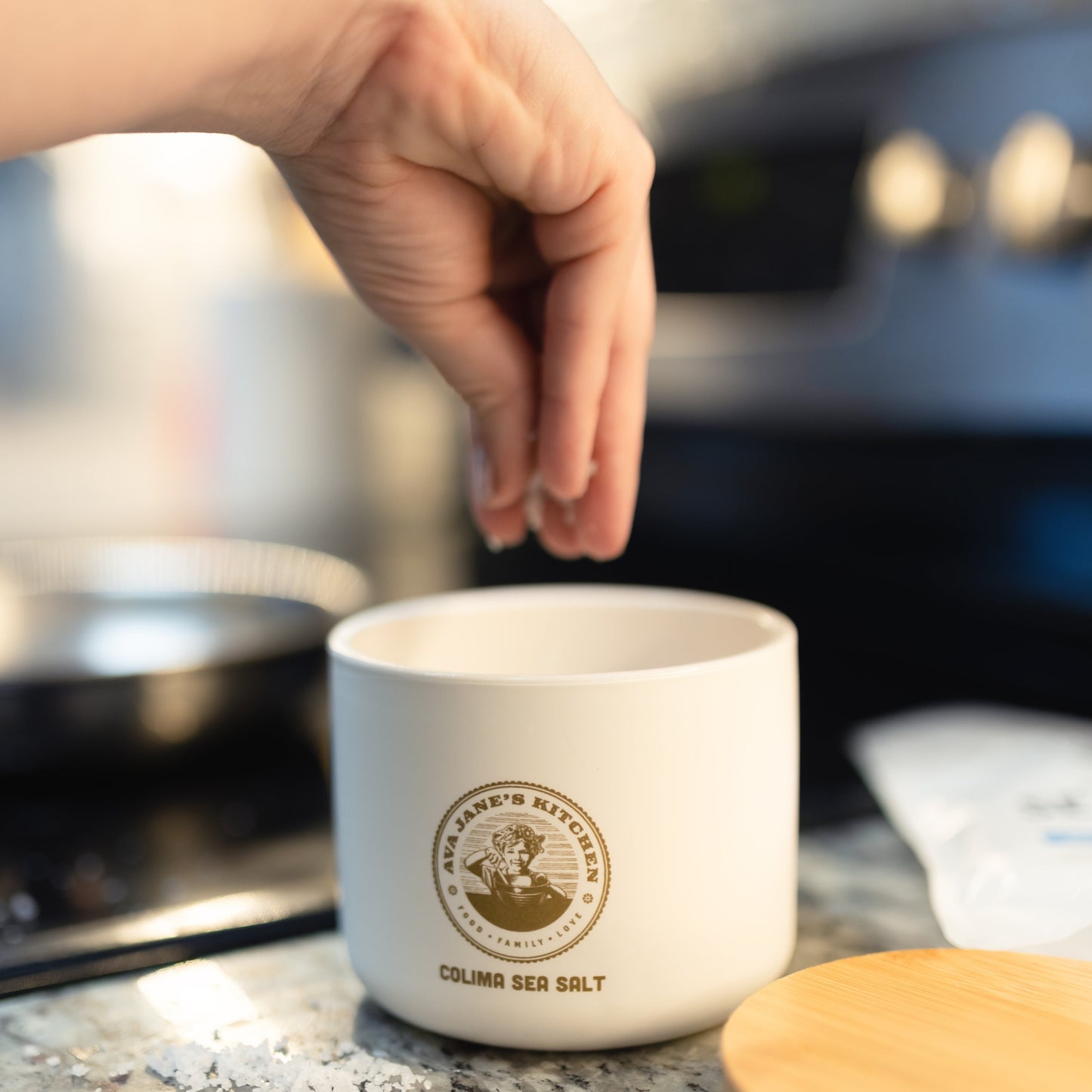 A person pinching Ava Jane Kitchen's Colima Sea Salt from the Porcelain Salt Cellar next to the stove while cooking