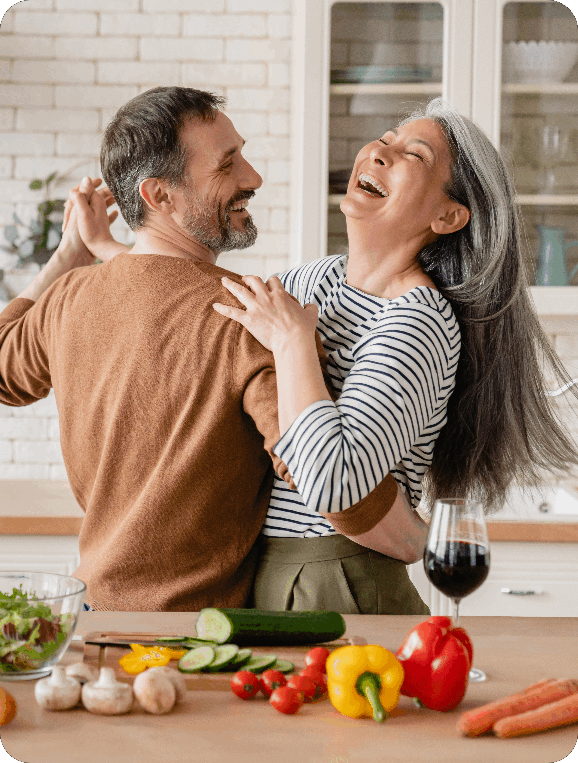 a couple dancing in the kitchen, while vegetables, sald bowl, and wine glass is present on kitchen island